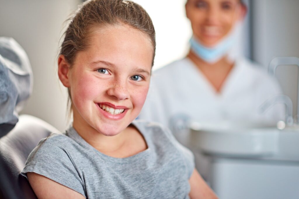 A young female patient smiles while sitting in a dental chair, during a myofunctional therapy consultation.